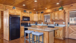 A kitchen with wooden cabinets, a black refrigerator, a microwave, a stove, and a sink. There are two bar stools and a window with blinds. - Heavenly Hideaway (Sevierville)