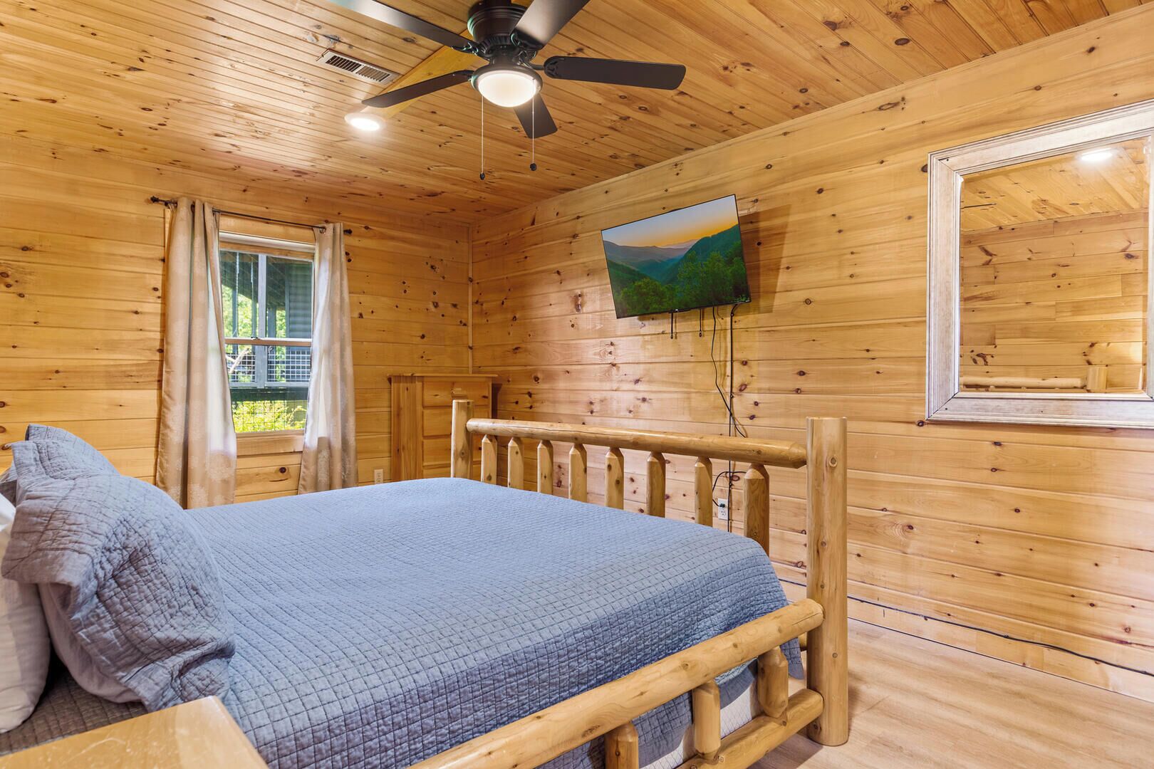 Habitación de cabaña de madera con cama, ventilador de techo, televisor montado en la pared y ventana con cortinas.