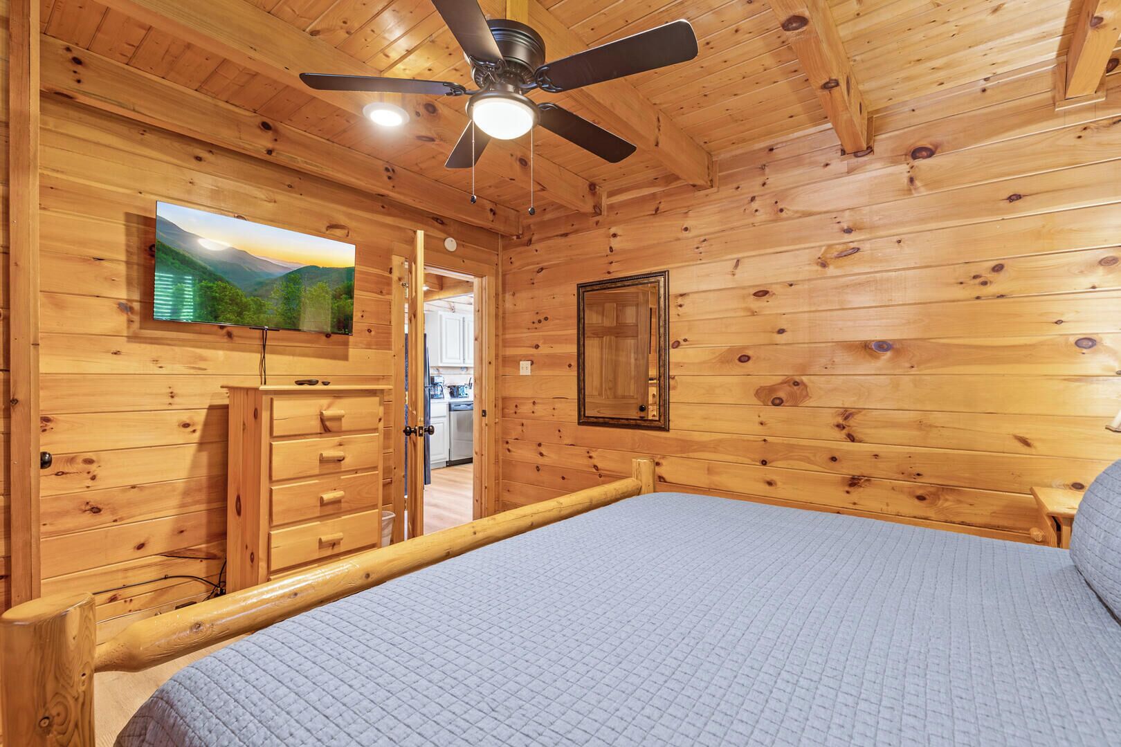 A wooden cabin bedroom with a bed, a ceiling fan, a TV, and a dresser.