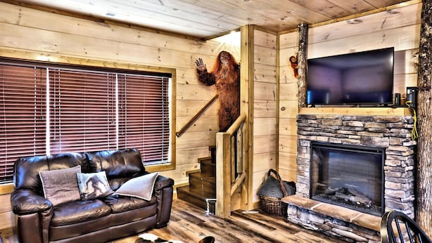 A cozy living room with a leather sofa, a fireplace, a TV mounted above it, and a wooden staircase.