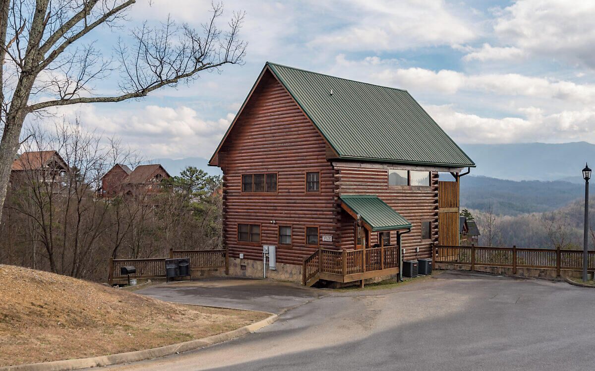 A log cabin with a green roof, a porch, and a view of mountains in the background.