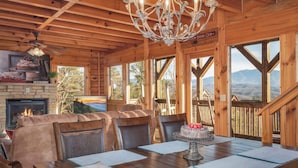 A wooden cabin interior with a dining table, chairs, a chandelier, a fireplace, and a view of the mountains. - Sweet Slice of Heaven (Sevierville)