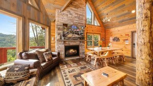 A cozy cabin interior with a fireplace, wooden furniture, and large windows offering a view of the outdoors.
