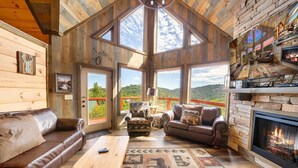 A cozy cabin interior with a fireplace, leather sofas, and large windows offering scenic views.