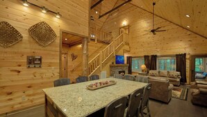 A cozy log cabin interior with a stone fireplace, a dining area with granite countertops, and a living room with a sofa and a television. - Adventure Lodge (Sevierville)