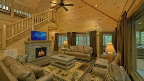 A log cabin interior with a stone fireplace, leather sofas, a television, and a staircase. - Adventure Lodge (Sevierville)