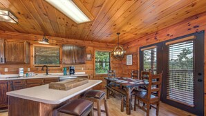 Interior rústico de una cabaña de madera con cocina y comedor, que incluye armarios de madera, una isla grande y una mesa de comedor con sillas.