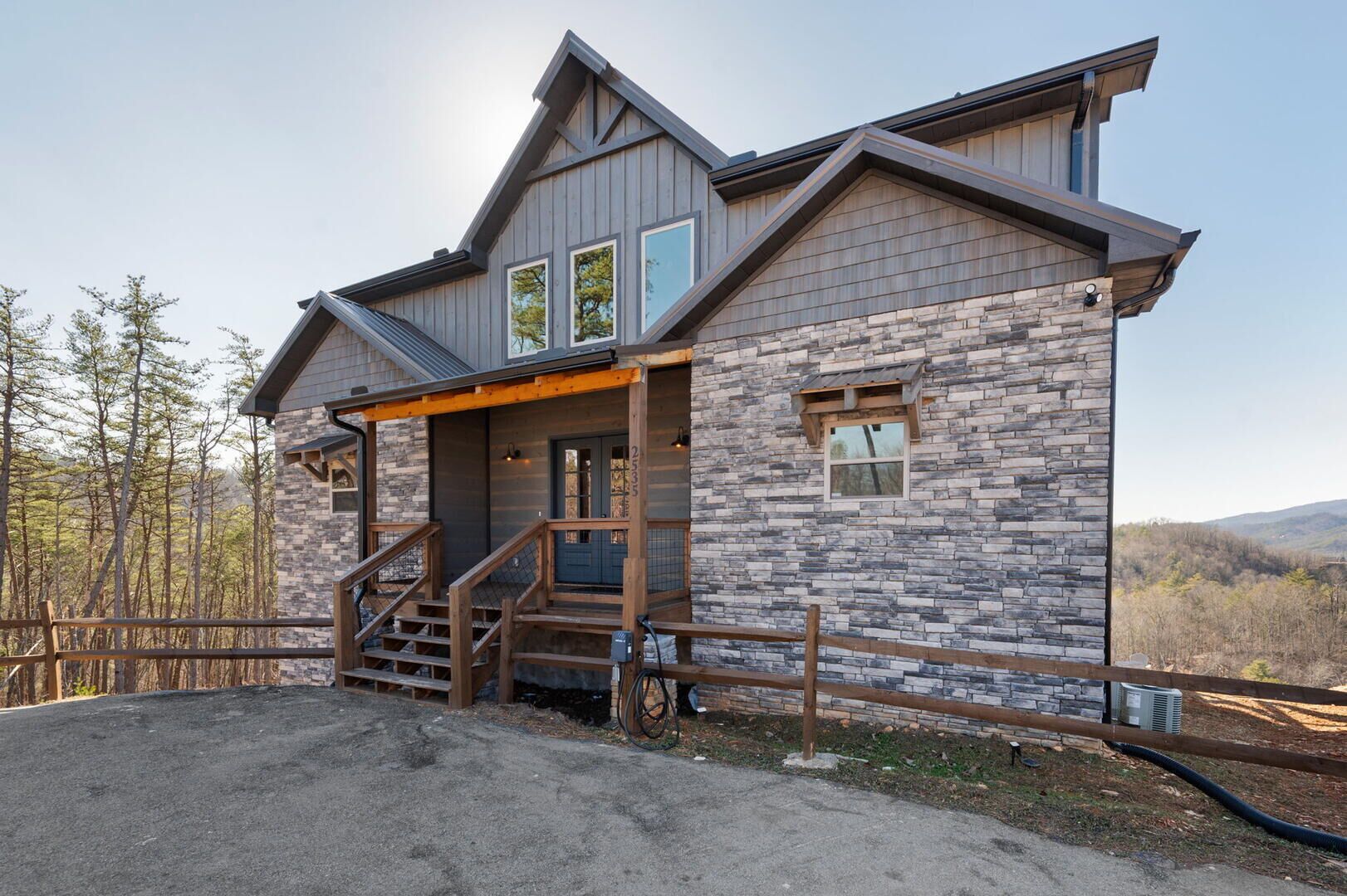 A modern two-story house with a stone facade, a wooden porch, and a view of the surrounding forest.