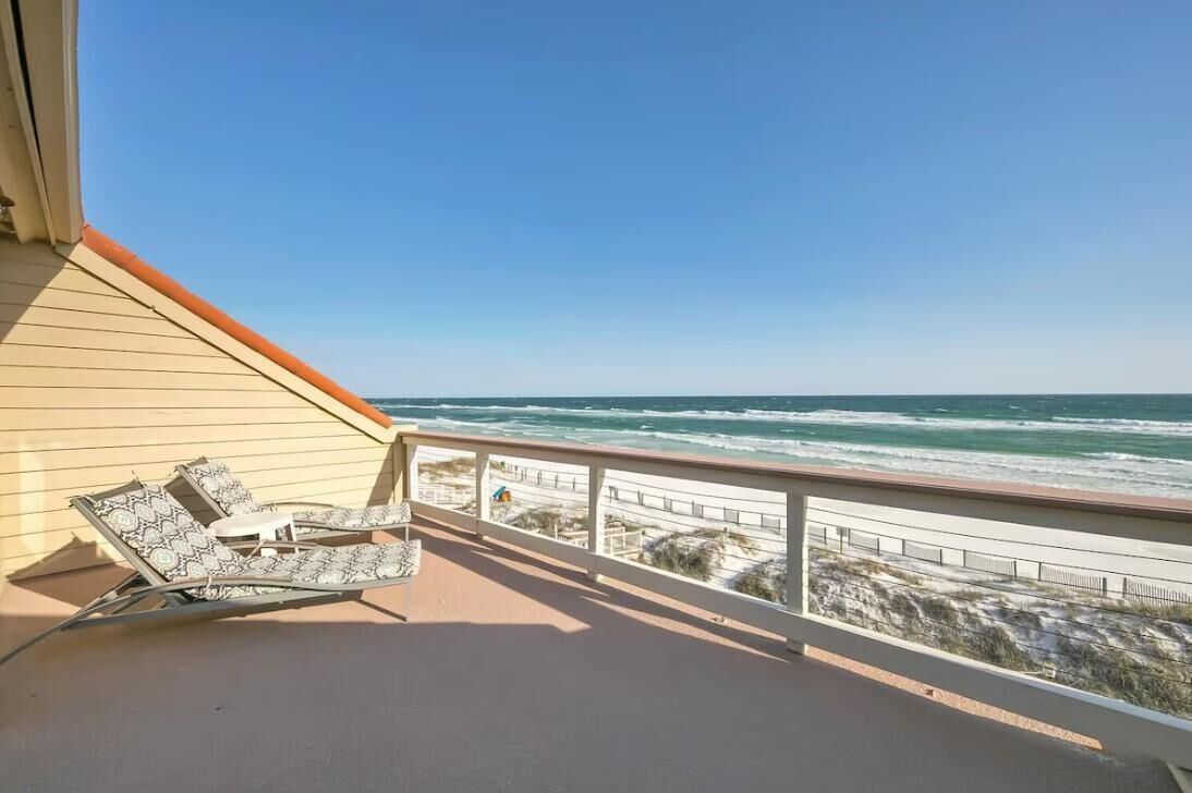 A balcony with a view of the ocean, a beach, and a chair.