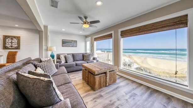 A spacious living room with a large gray sectional sofa, a woven coffee table, and a ceiling fan. A large window offers a view of the beach. - Green Reef #6- Beachfront Bliss (Destin)