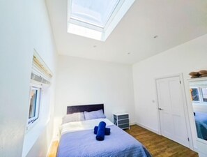 A bedroom with a skylight, a bed with a blue blanket, a small bedside table, and a wardrobe. - Orpington Cozy Retreat With 3 Bed (Orpington)