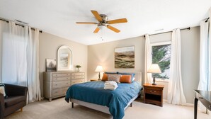 Une chambre à coucher comprenant un lit, une commode, une table de chevet, une chaise, un ventilateur de plafond et une fenêtre avec des rideaux.