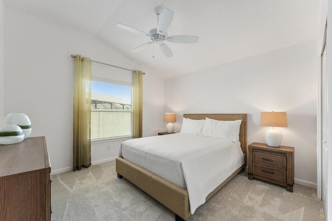 Un dormitorio con cama, ventilador de techo, mesita de noche con lámpara, ventana con cortinas y vista a un puente.