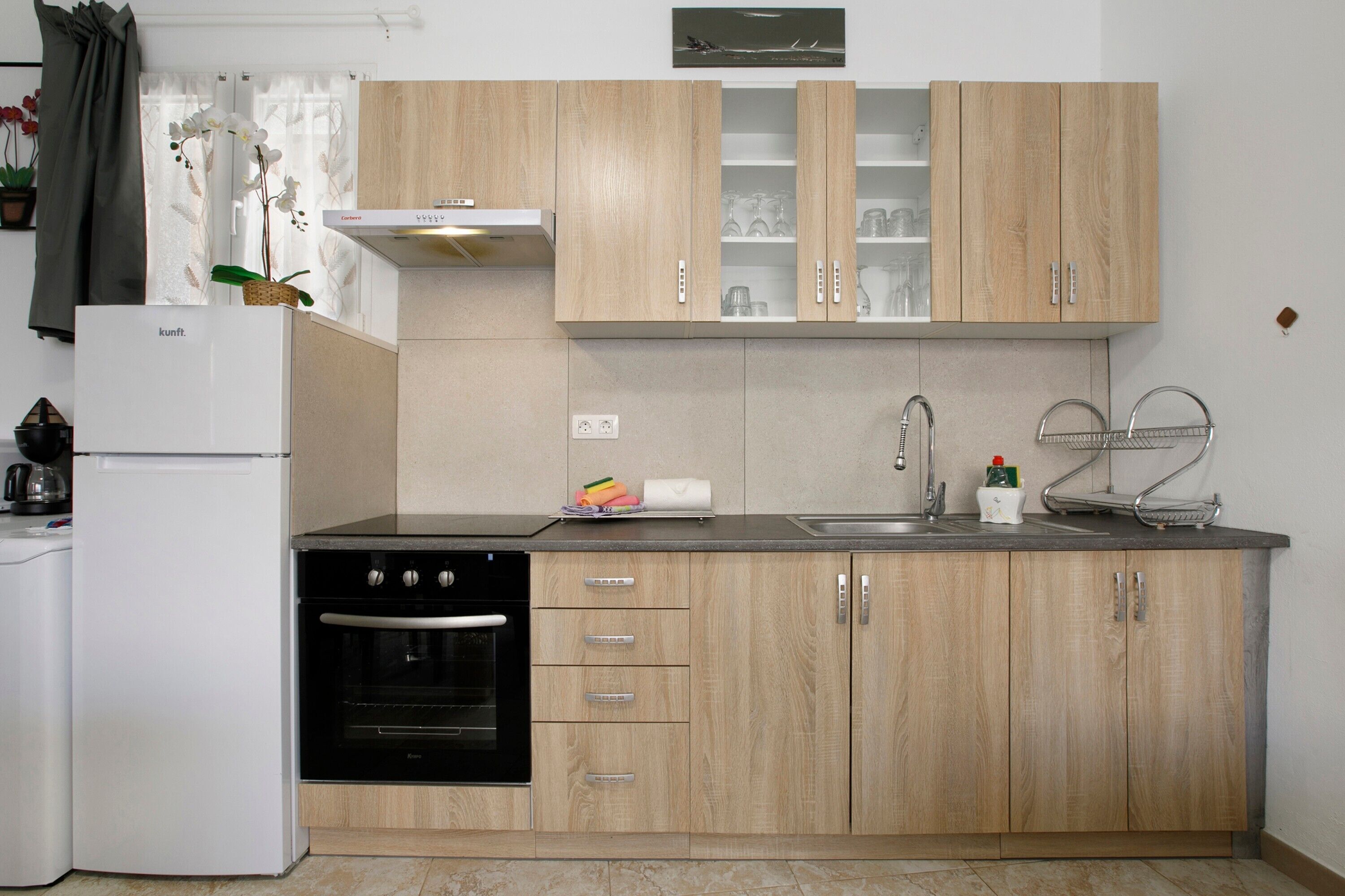 A kitchen with wooden cabinets, a built-in oven, a refrigerator, a sink, and a window with a curtain.