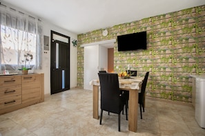 A dining area with a wooden table, chairs, a TV mounted on the wall, and a kitchen area with cabinets and a refrigerator.
