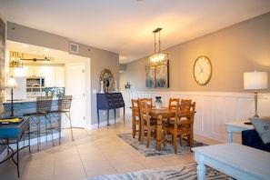 A dining area with a wooden table and chairs, a chandelier, a clock, and a kitchen in the background.