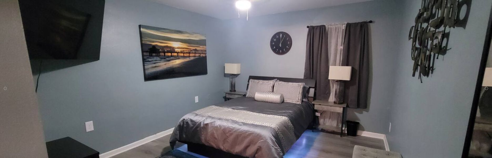 A bedroom with a bed, a clock, a painting, and a TV on the wall.