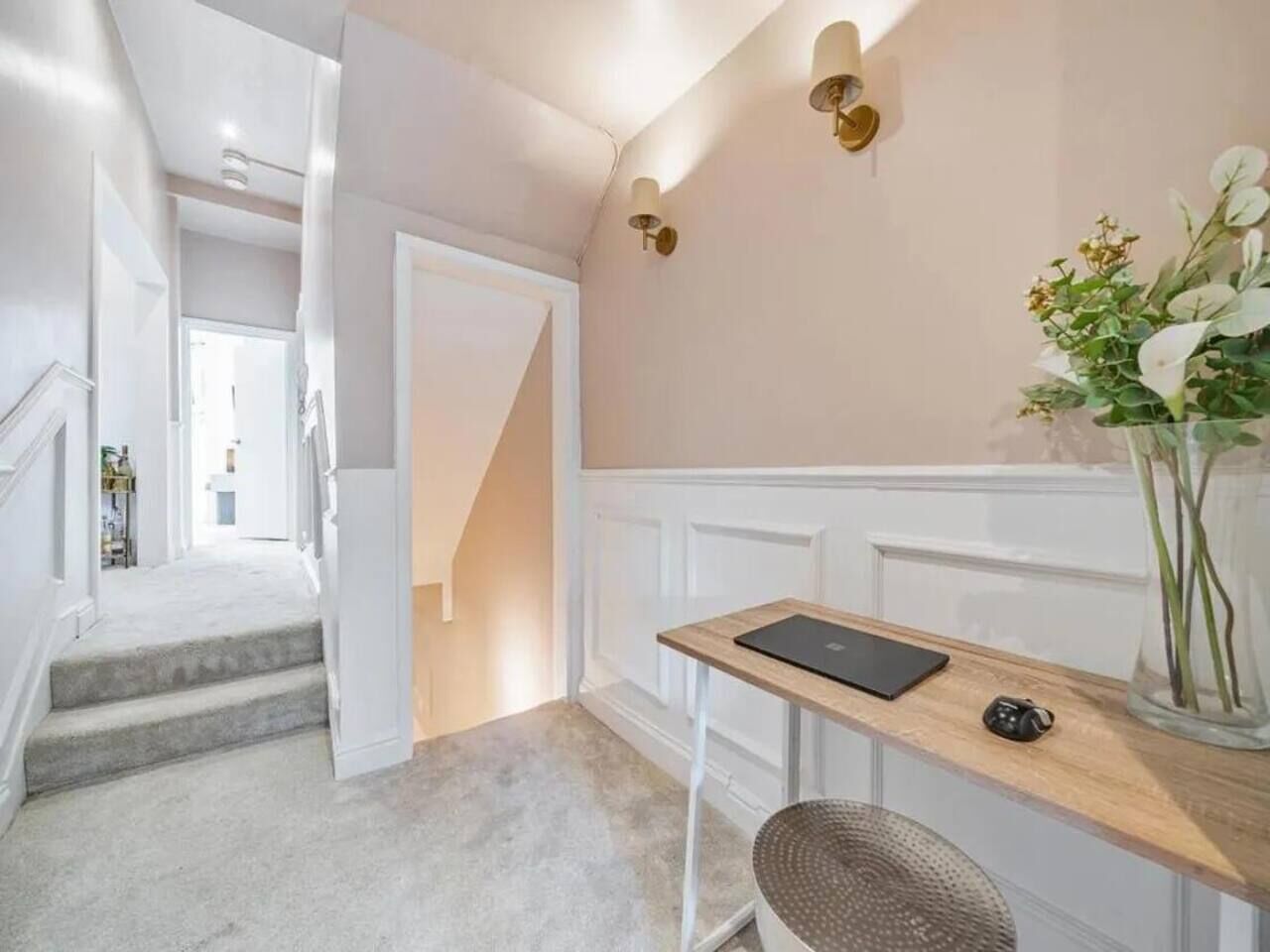 A hallway with a staircase leading to another room, a small desk with a laptop, and a vase with flowers.