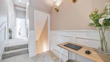 A hallway with a staircase leading to another room, a small desk with a laptop, and a vase with flowers.