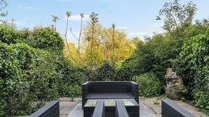Une terrasse de jardin avec un canapé en osier et une table à plateau de verre, le tout entouré d’un feuillage dense et d’arbres.