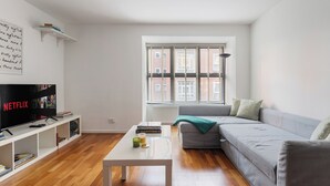A modern living room with a flat-screen TV, a white coffee table, a grey sofa, and a window with blinds. - Elegant 1BR Flat in Westminster Near Victoria Tube (London)