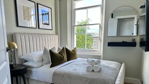 A neatly arranged bedroom with a large bed, a bedside table with a lamp, a window with a view, and a mirror on the wall. - Modern 1BR Flat in Peaceful North Kensington (London)