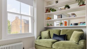 A green sofa with two cushions, a white radiator, and a window with a view of brick buildings. - Modern 1BR Flat in Peaceful North Kensington (London)