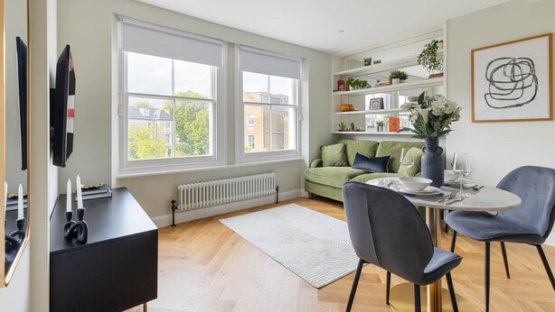 A modern living room with a dining area, large windows, a green sofa, and a white rug. - Modern 1BR Flat in Peaceful North Kensington (London)
