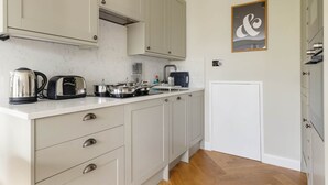 A kitchen with white cabinets, a countertop with a kettle, toaster, and pots. There is a door with a handle and a wall-mounted artwork. - Modern 1BR Flat in Peaceful North Kensington (London)