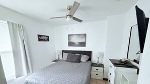Ein Schlafzimmer mit Bett, Nachttischen, einem Schreibtisch, einem Spiegel, einem Deckenventilator und einem Fenster mit Vorhängen.