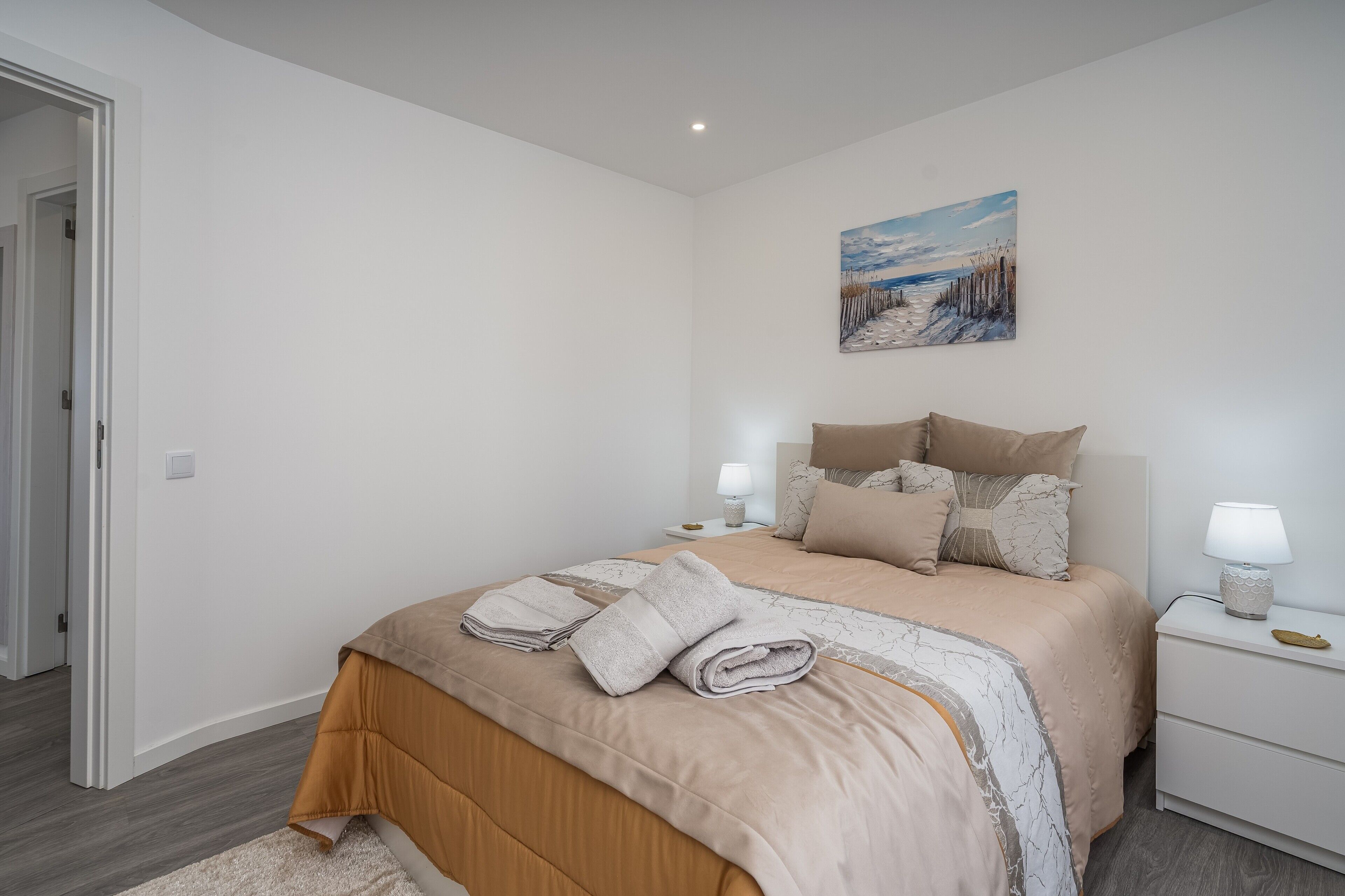 A bedroom with a bed, pillows, a nightstand with a lamp, a painting on the wall, and a door.