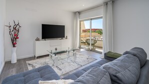 Una sala de estar moderna con una televisión de pantalla plana, una mesa de centro de vidrio, un sofá y vistas al exterior.