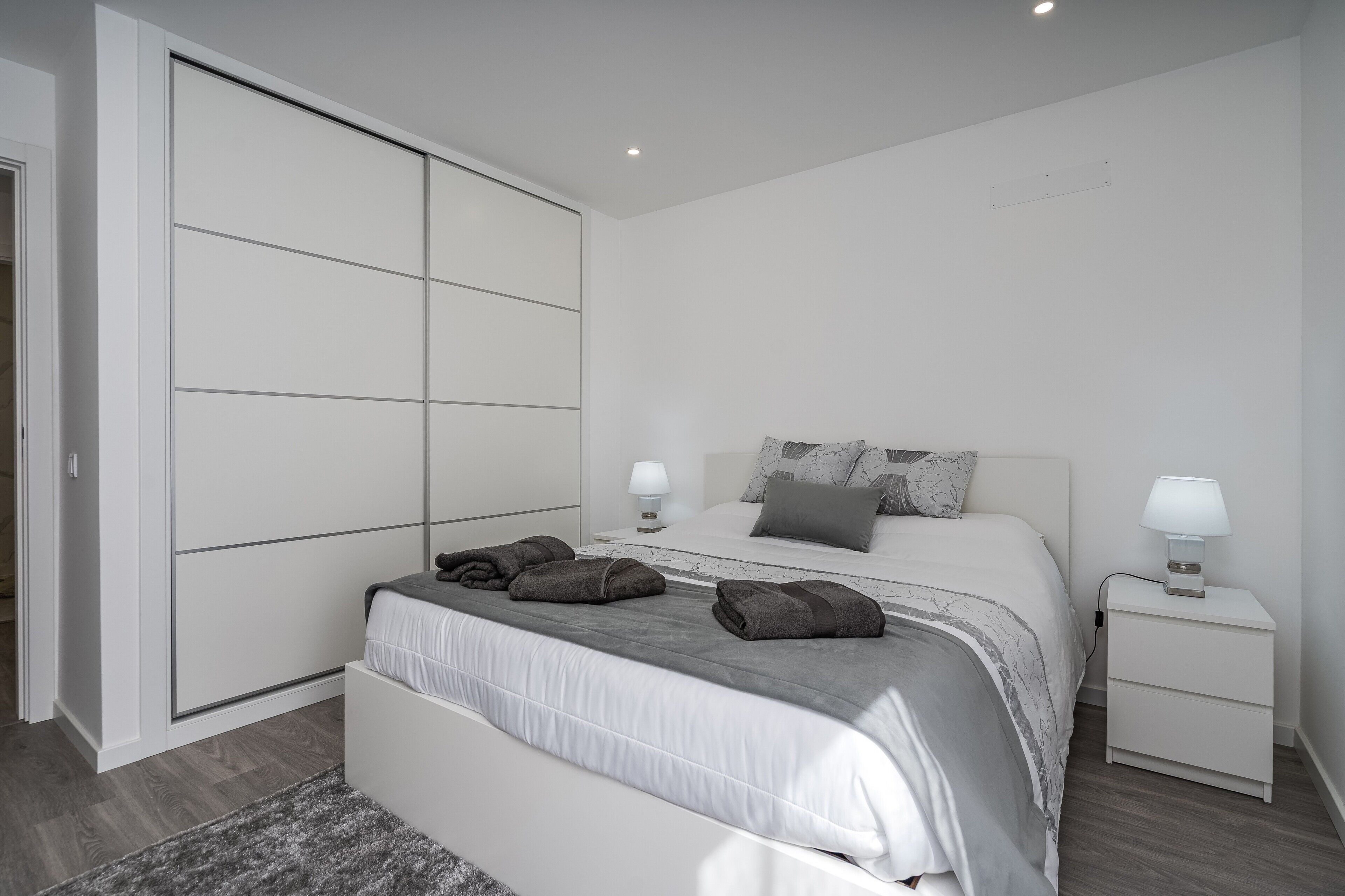 A modern bedroom with a large wardrobe, a bed with a grey headboard, and bedside tables with lamps.