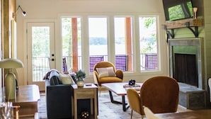 A spacious living room with a high ceiling, large windows, a fireplace, and a television mounted on the wall.