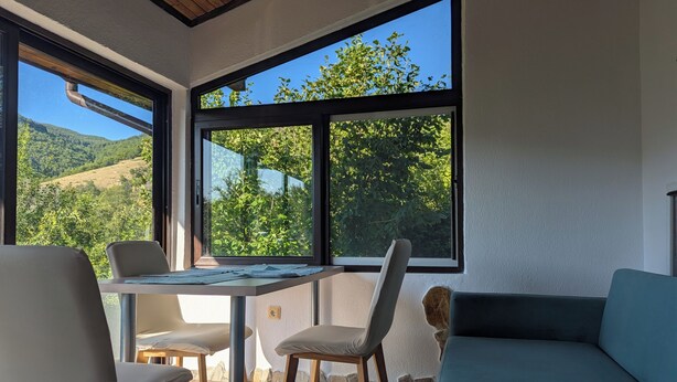 Ein Zimmer mit Tisch und Stühlen, einer Couch und großen Fenstern mit Blick auf Grünflächen und Hügel.