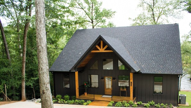Une cabane dotée d’un toit à pignon très incliné, de grandes fenêtres et d’un porche à l’avant avec une balustrade.