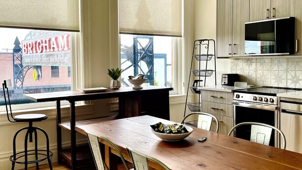 A modern kitchen with a wooden table, metal chairs, and a view of a cityscape through large windows. - Stunning new loft in historic Union Block. Brand new, walk to everything. (Brigham City)