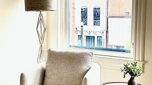 A room with a beige armchair, a wooden side table with a vase, a floor lamp, and a view of a building through a window. - Stunning new loft in historic Union Block. Brand new, walk to everything. (Brigham City)