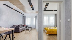 Ein modernes Wohnzimmer mit einer Couch, einem Couchtisch und einem Esstisch mit Stühlen. Im angrenzenden Raum befindet sich ein Bett mit gelber Bettdecke.