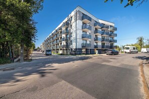 Un edificio moderno de varios pisos con balcones, una calle al frente y vehículos estacionados cerca.