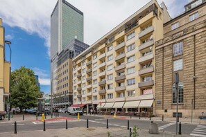 Un hotel de varios pisos con fachada moderna, un llamativo letrero de Samsung y vista a la calle con un paso de cebra y bolardos.