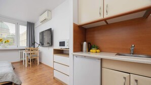A compact room with a bed, a desk, a chair, a small kitchenette, and a window with a view of a building.