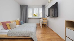 A bedroom with a bed, a desk, a chair, a TV, and a window with curtains.