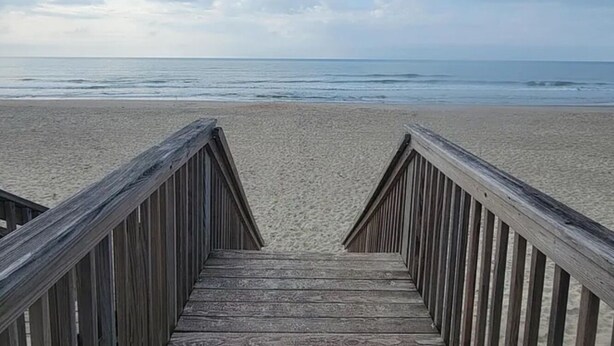 Een houten looppad dat naar een zandstrand leidt, met een kalme zee en een gedeeltelijk bewolkte lucht.