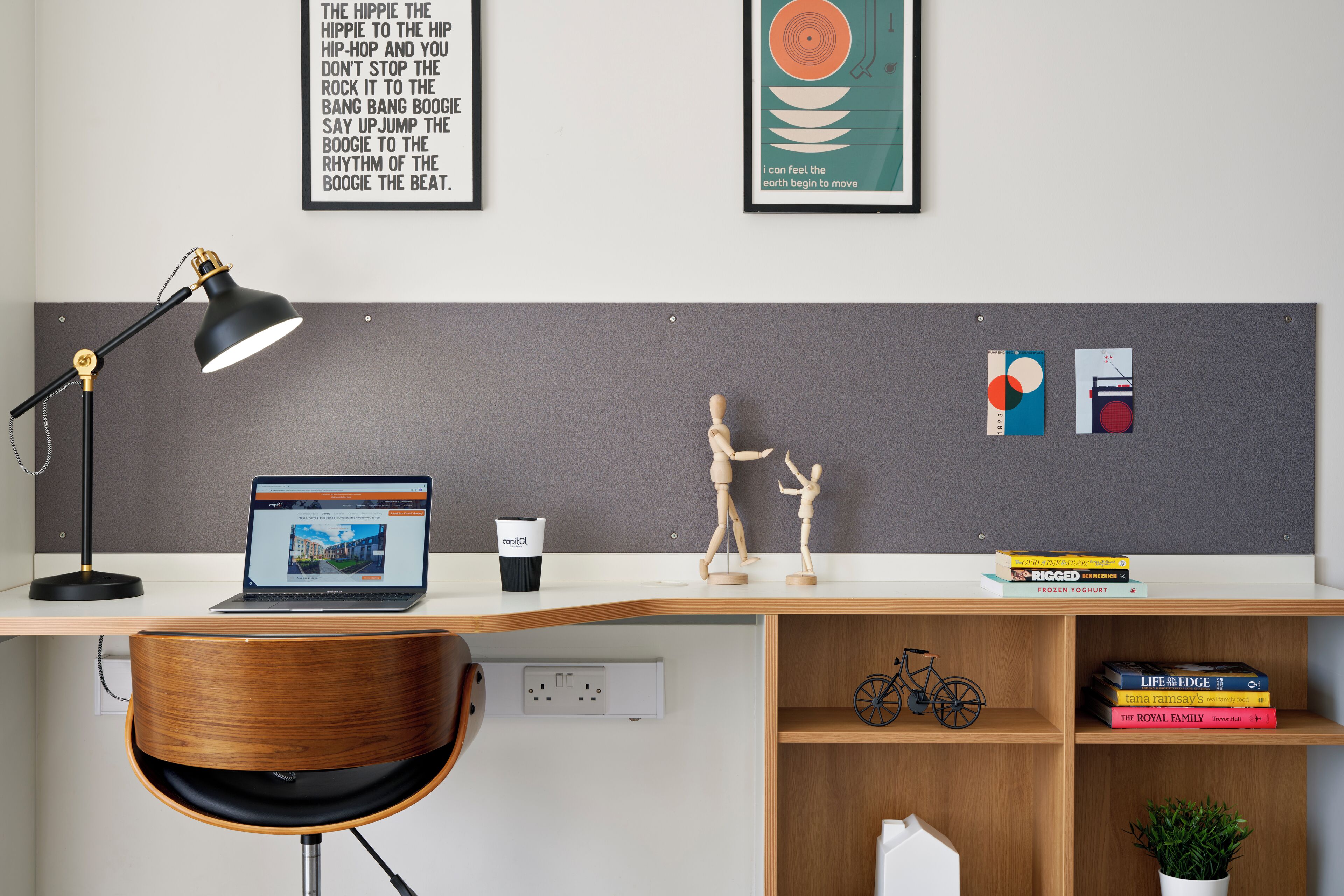 A modern workspace with a laptop, desk lamp, and wooden chair. There are books, a coffee cup, and a small plant on the desk.