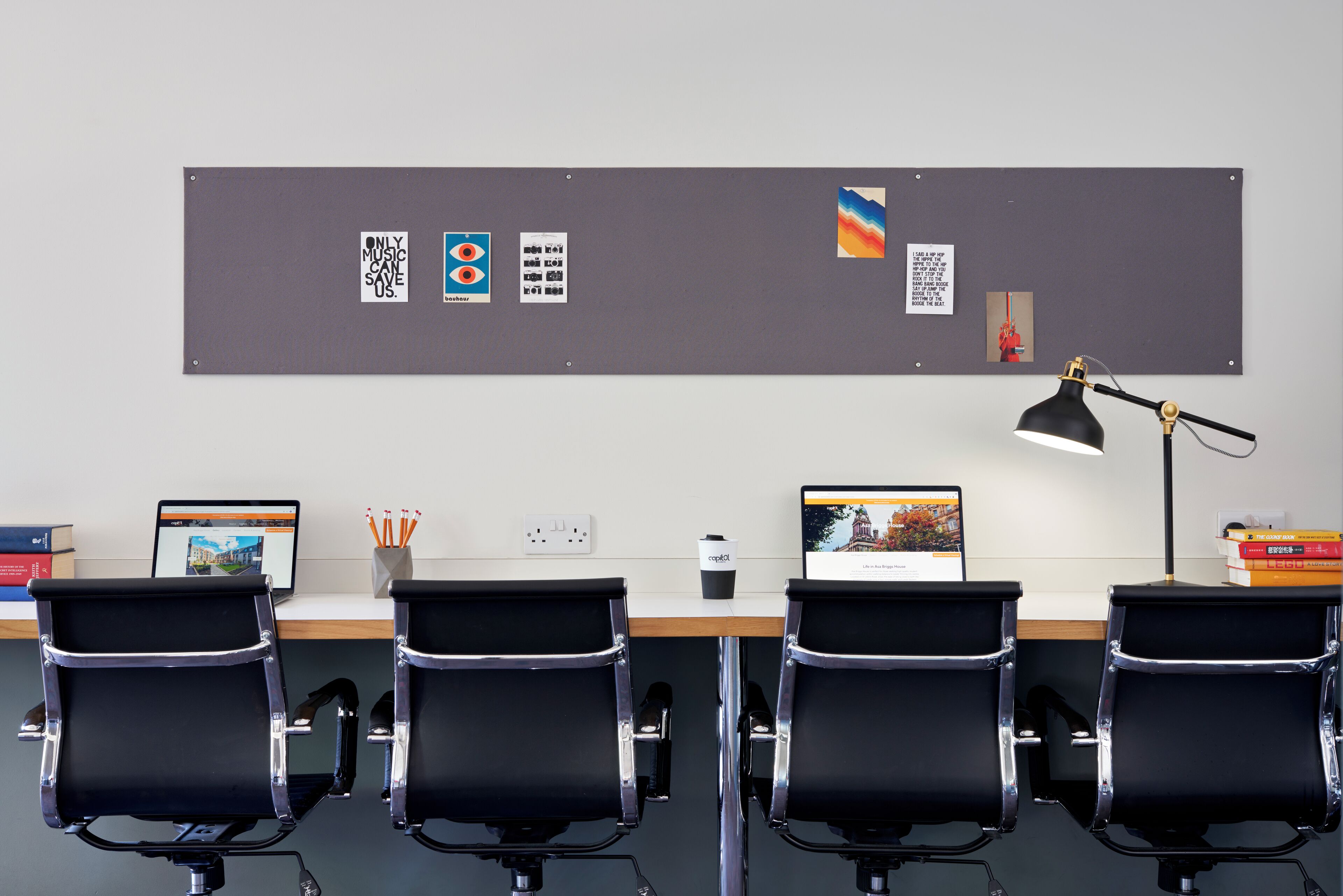 A modern office with four black office chairs, two laptops, a desk lamp, and a bulletin board with various items pinned to it.