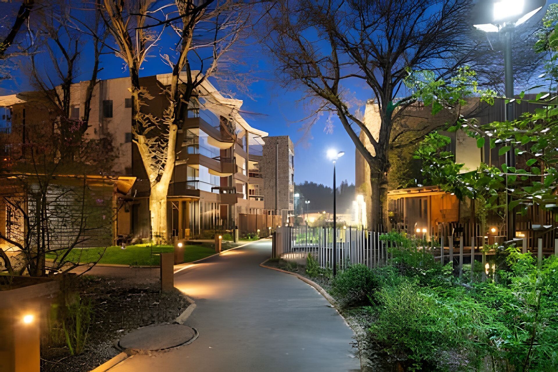 Vue nocturne d’un quartier résidentiel moderne, avec un chemin sinueux, des arbres et des lampadaires illuminés.