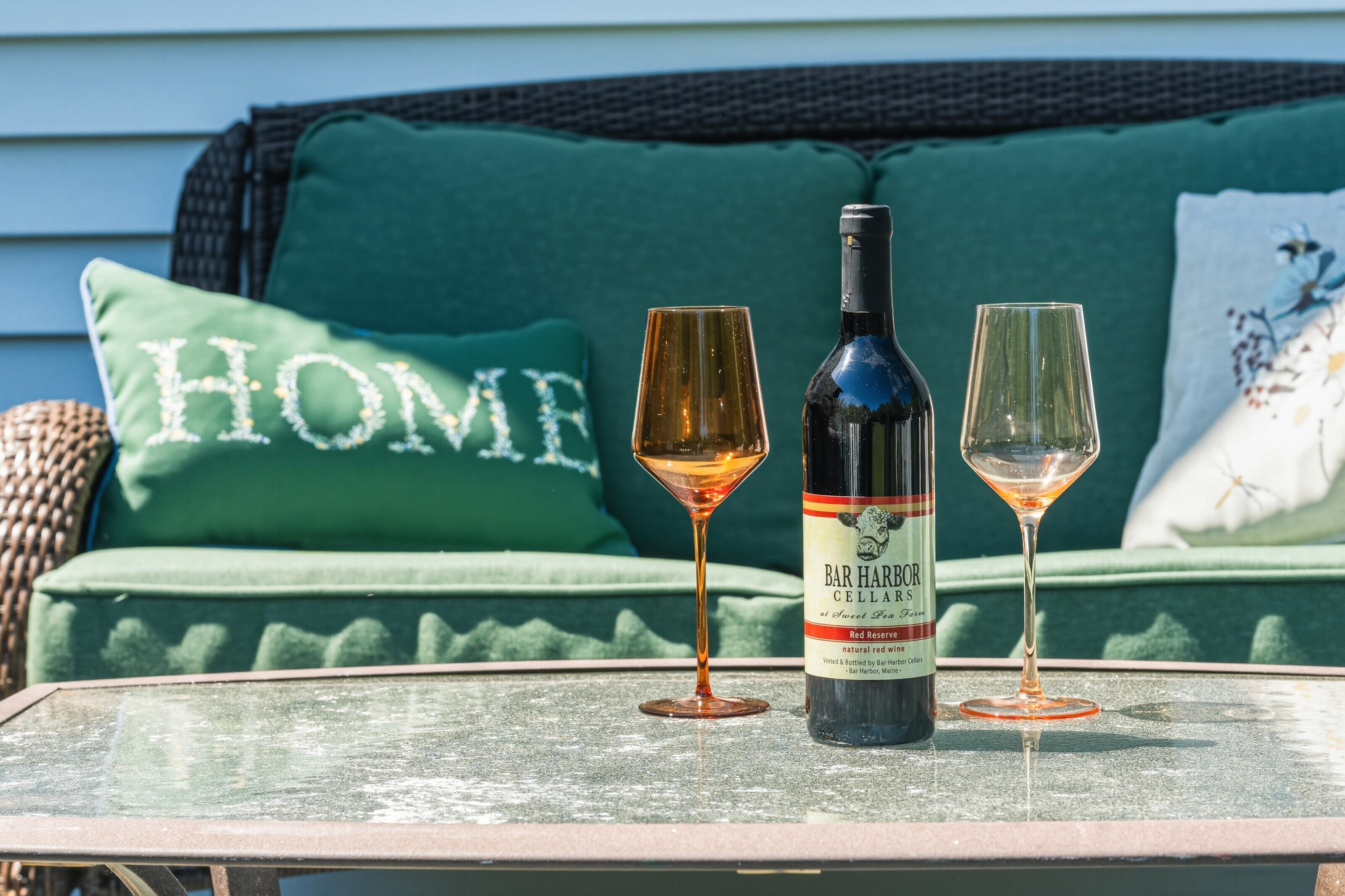 A bottle of Bar Harbor Cellars red reserve wine and two glasses on a table with a green cushioned sofa in the background.