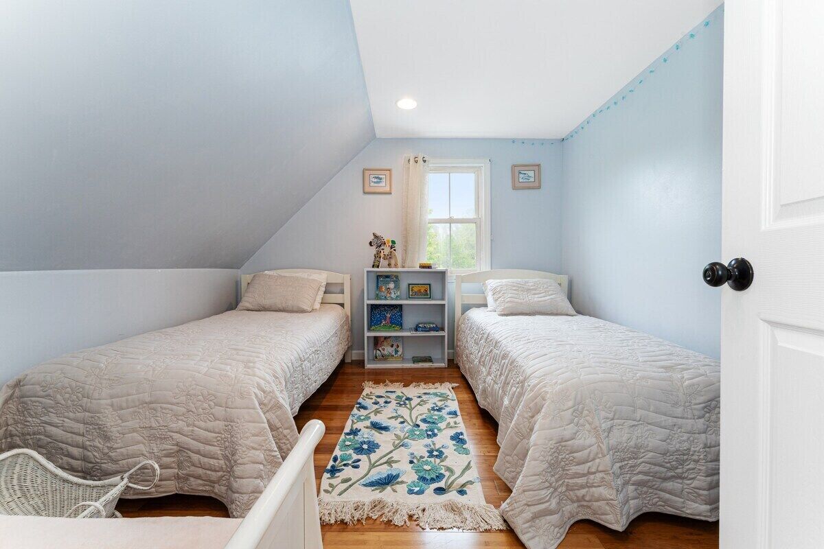 Un dormitorio con dos camas, una ventana, una estantería con libros y una alfombra estampada con motivos florales.
