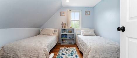 Un dormitorio con dos camas, una ventana, una estantería con libros y una alfombra estampada con motivos florales.